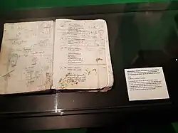 Dans une vitrine, un livret de scénario en mauvais état, ouvert à une page agrémentée de dessins et notes. Des tâches vertes en bas de la page droite.