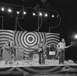 Photo en noir et blanc d'un groupe de musiciens sur une scène. On peut voir un joueur de mandoline, un guitariste et un bassiste, tous en train de jouer et de chanter dans des micros sur pied. Derrière eux, une batterie est visible, mais pas le batteur qui en joue.