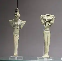 Deux statuettes féminines au corps effilé et aux épaules larges. Le crâne de celle de gauche est pointu et recouvert de matière sombre rappelant sans doute une chevelure. Elles sont vues de face, le fond est gris bleuté et il manque la tête de la statuette de droite.