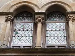 Photo couleur d'une fenêtre en pierre double à arc gothique encadré de colonnettes. La baie est munie de vitraux en verres en losange.