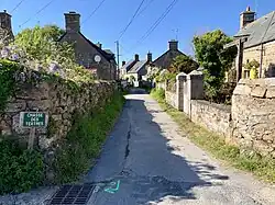 Photographie en couleurs prise à l'entrée d'une voie de circulation. Des maisons à gauche et à droite sont séparées de la voie par d'épais murs en pierre.