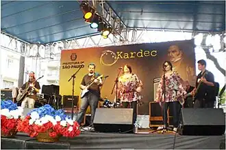 Festival spirite dans les rues de São Paulo en 2010. L'image représente une scène musicale surmonté d'un toit bleu. Des chanteurs et musiciens travaillent devant un panneau géant présentant le portrait d'Allan Kardec et le logo de la municipalité de Sao Paulo.
