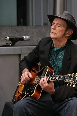 Photo d'un homme blanc d'un certain âge portant un chapeau noir et une veste de la même couleur, avec une chemise verte. Assis devant un micro, il joue de la guitare les yeux fermés.