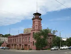 Photographie en couleur montrant un bâtiment couleur rouge-rose, avec sa tour centrale, coiffée d'une petite toiture pointue. Des fenêtres rectangulaires ornent les façades, tandis que des véhicules sont stationnés devant.