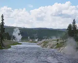 Photographie de la rivière Firehole et d'un geyser en éruption en arrière-plan.