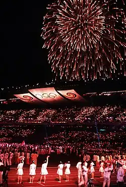 Photographie de l'intérieur d'un stade, de nuit. Des athlètes sont regroupés sur le terrain central, et dans le ciel est tiré un feu d'artifice.