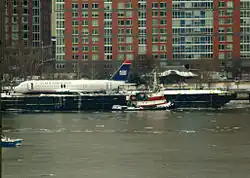 Photo montrant l'avion endommagé, sur la gauche de l'image, après sa récupération du fleuve gelée, au premier plan. L'avion se trouve sur la berge, avec la ville en arrière-plan.