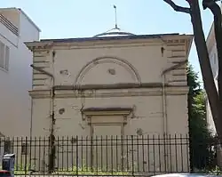 Façade du cabinet d'Histoire Naturelle. Façade sobre dont le crépi blanc sale est fissuré et s'écaille. Des mauvaises herbes tout autour.