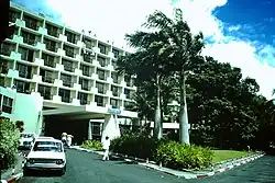 Bâtiment de plusieurs étages, sur une rue ensoleillée bordée de palmiers.