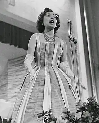 Image en noir et blanc dune femme début au centre, chevelure frisée, bouche ouverte devant un micro sur pied, bras écartés, portant gants longs blancs et robe évasée sur le bas, avec rainures verticales inégales blanches et noires.