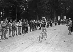 Photographie en noir et blanc montrant un cycliste lors d'une course avec des spectateurs sur le bord de la route.