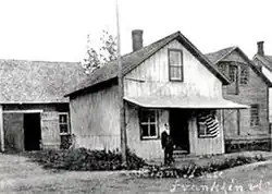 Une cabane revêtue de tôle devant laquelle se tient une personne habillée en uniforme.