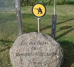 Boule de pierre portant inscription. «&nbsp;Für die Opfer der Unmenschlichkeit&nbsp;». À l'arrière se trouve une section de clôture et un panneau jaune montrant un soldat agenouillé épaulant son arme.