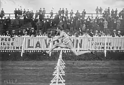 Photographie en noir et blanc d'un coureur à pied qui saute une haie dans un stade