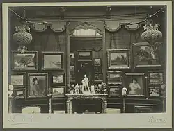 Vue d'une cimaise de la galerie Henry Vasnier sur laquelle sont accrochés des tableaux, d'autres sont posés à même le sol, contre elle. Au centre se trouve une cheminée surmontée d'un miroir dans lequel se reflète le photographe. Des sculptures et des objets d'art sont disposées sur sa tablette. D'autres sculptures sont posées sur des colonnes.
