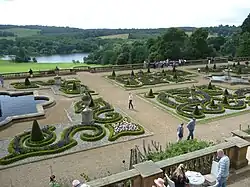 Photo en plongée sur un jardin avec des parterres géométriques et des bassins, avec un lac et des bosquets à l'arrière-plan