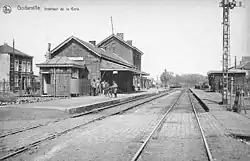Carte postale en noir et blanc représentant une gare, des voies ferrées et quelques personnes.