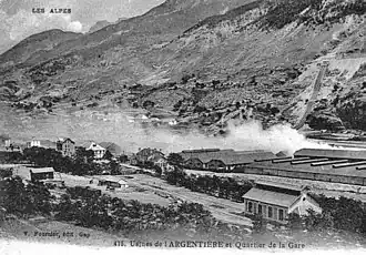 Photo de bâtiments industriels bas enfumés au pied d'une montagne.