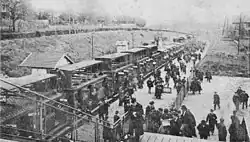 Photo noir et blanc prise depuis une passerelle, montrant un train de banlieue d'où descend une foule de voyageurs dont certains lisent des journaux.
