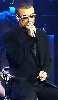 Photographie couleur d'un homme assis qui porte de la main droite un micro à la bouche.