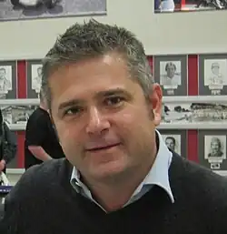 Photographie d'un homme blanc, vu de face, inclinant sa tête, cheveux gris, souriant.