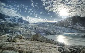 Photographie en couleurs de roches brunes et grises, aux formes arrondies, bordées par les eaux d'un fjord, un glacier visible en arrière-plan.