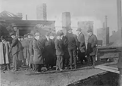 Photo en noir et blanc. Une vingtaine d'hommes, en pardessus et chapeaux melon, examinent les dégâts de l'incendie sur le toit de l'hôtel. On distingue un morceau de bois brûlé au pieds des hommes au premier plan, et des cheminées en brique à l'arrière plan.