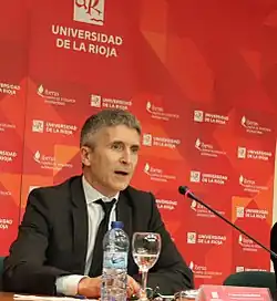 Photo d'un homme auc heveux gris en costume parlant dans un micro, à l'arrière un fond rouge