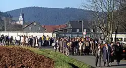 Un grand nombre de personnes de tous ages se trouvent le long d'une route en face d'un mur de béton derrière lequel se trouvent des maisons et une église.