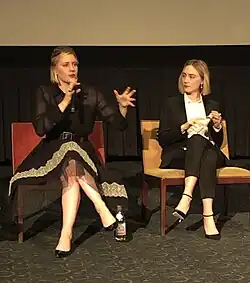 Photographie de deux femmes habillées en noir et assises devant un écran de projection. Celle de gauche, habillée en robe, tient un micro et est en train de parler, tandis que celle de droite la regarde.