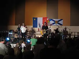 Photographie du groupe de musique Suroît durant l'un de ses spectacles. Sur le mur au fond de la scène se trouvent le drapeau de l'Acadie et celui de la Nouvelle-Écosse.