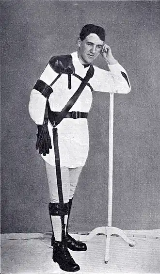 Photographie en noir et blanc d'un homme debout muni d'un appareillage orthopédique complexe, prenant la jambe droite.