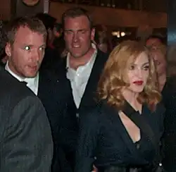 Un homme en costume accompagne une femme blonde avec une robe noire et le bras gauche en écharpe