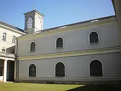 Façade blanche sur deux niveaux, présentant six ouvertures cintrées. En haut à gauche, le clocher.