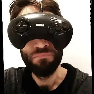 Portrait d'un homme brun barbu, qui met une manette de jeu vidéo devant son visage comme si c'était une paire de lunettes.