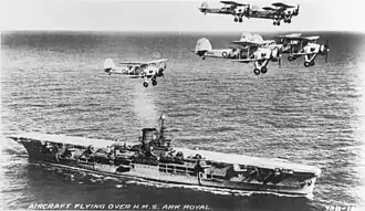 Photographie en noir et blanc d'un porte-avions vu de tribord, survolé par des avions biplans.