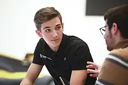 Photographie d'un jeune homme, vu de trois-quarts droit, blanc, châtain, aux yeux marrons, au polo noir, écoutant quelqu'un de dos.