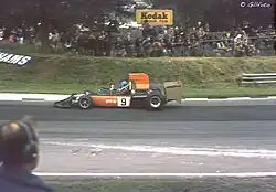 Photographie d'une monoplace noire et orange, vue de profil, sur une piste. En arrière-plan, des dizaines de spectateurs