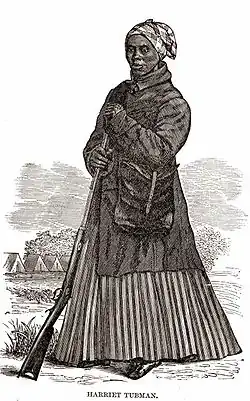 Une femme noire, en pied, appuyée sur un fusil. Elle est coiffée d'un turban et porte un grand sac en bandoulière