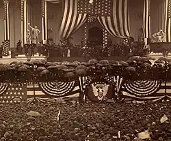 Photographie d'une estrade décorée par des drapeaux américains. De nombreux participants tiennent des parapluies et toutes les personnes de la foule en contrebas portent des chapeaux.