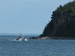 Un petit navire de pêche navigue sur l'eau à proximité d'une falaise couverte d'arbres.