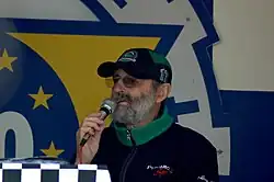 photo d'un homme, habillé en noir et vert, parlant dans un micro, de face, trois-quart