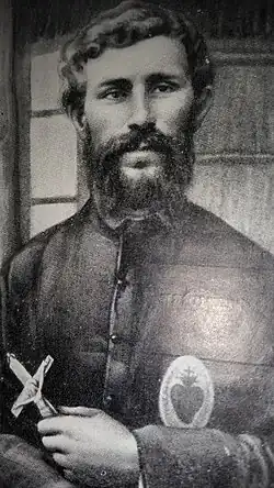 Photo d'un prêtre, barbu, un Sacré-Cœur sur la poitrine et un crucifix à la main, devant une cloison