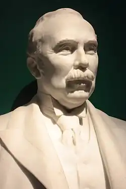 Photo du haut d'une statue d'un homme, incluant le buste. Le visage est détaillé, en particulier au niveau des yeux ; il porte une moustache et un costume cravate.