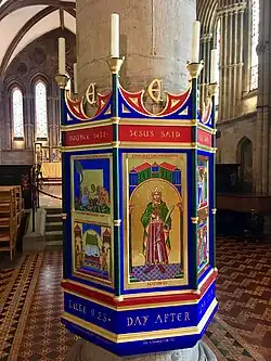 Photo prise à l'intérieur d'une église montrant des panneaux décorés d'images aux couleurs vives, exposés autour d'un pilier de pierre. L'image qui fait face à l'objectif présente, dans un style médiévalisant, la figure d'un homme debout portant une couronne et tenant dans sa main une église en modèle réduit. La légende indique : Ethelbert King and Martyr