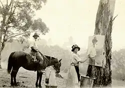 Photographie en noir et blanc. Au premier plan, l'artiste regarde le photographe tout en ayant à la main un pinceau à hauteur de la toile qui est suspendue à un arbre. Au deuxième plan, un homme à cheval de profil. En arrière plan, le bush australine.