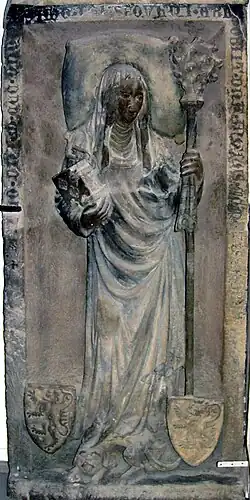 Photographie d'une épitaphe en relief montrant une femme qui tient une crosse et aux pieds de laquelle deux blasons sont posés.