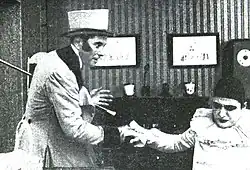 Un homme en redingote, gibus et canne tient la main d'un Pierrot agenouillé et contrit, tout en le regardant.