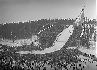 Photographie en noir et blanc d'un tremplin de saut à ski. Une grande foule est présente autour et en bas du tremplin.