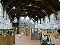 Photographie de l'intérieur d'un large bâtiment rectangulaire en pierre. La charpente sombre est apparente.